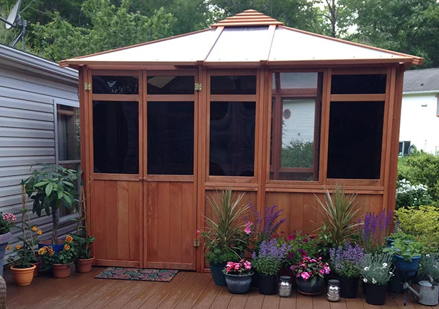 Wooden gazebo with dark windows on a deck, surrounded by assorted potted plants and greenery.