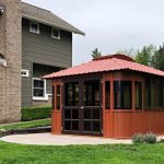 The Aspen 12x14, a red rectangular hot tub gazebo with a metal roof and large windows, sits on a circular concrete patio in a grassy backyard beside a two-story house, with trees and other houses visible in the background.
