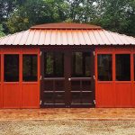 Red wooden gazebo with a gabled roof, centered in a garden setting surrounded by trees and bushes.