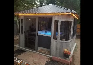 Gazebo With Illuminated Roof And Glowing Hot Tub Inside, Surrounded By Trees And Garden Elements.