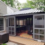 Wooden deck with a gray gazebo featuring large glass doors and outdoor dining set, surrounded by trees and a fence.