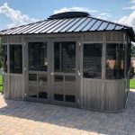 A small, modern gazebo with glass windows and a metal roof, situated on a patio surrounded by grass and trees.