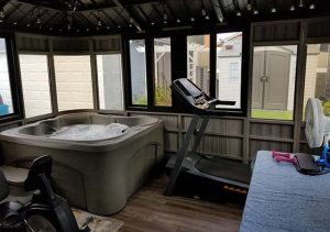 Indoor Room With A Jacuzzi, Treadmill, And Exercise Bike With Windows Showing An Outdoor Shed.