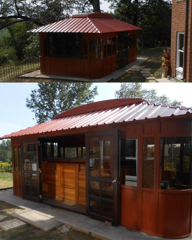 Wooden gazebo with a sloped red metal roof and glass doors, situated outdoors with trees in the background.