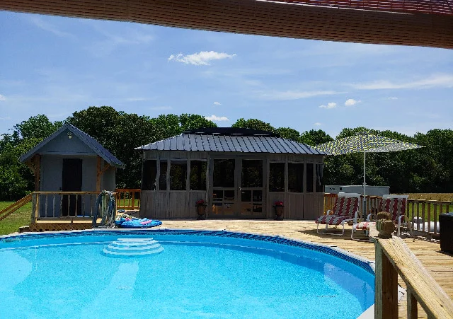 Backyard with a round pool, small gazebo, chairs, and umbrella surrounded by trees under a blue sky.
