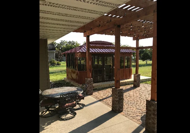 Patio with a round table set and wooden gazebo, under a pergola, in a sunny backyard setting.
