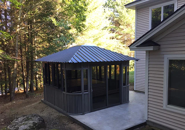 Outdoor gazebo with metal roof attached to a house on a concrete patio, surrounded by trees.