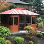 A wooden gazebo with a red metal roof in a landscaped garden with shrubs and trees around.