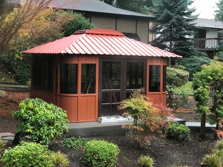 A wooden gazebo with a red metal roof in a landscaped garden with shrubs and trees around.