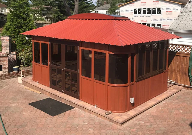 Wooden gazebo with windows and a red metal roof on a stone patio.