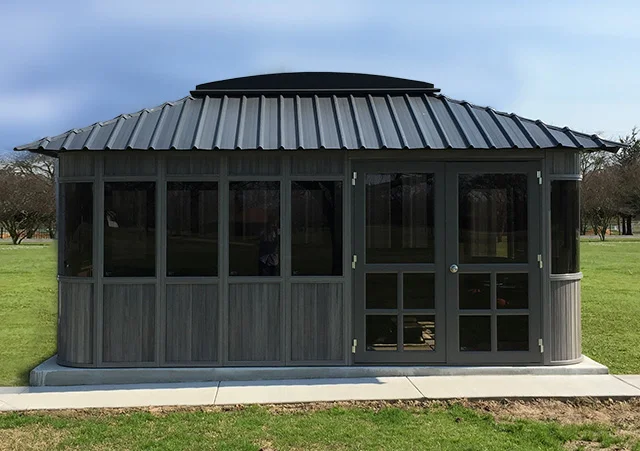 A small, grey gazebo with dark windows and a metal roof on a grassy area under a blue sky.