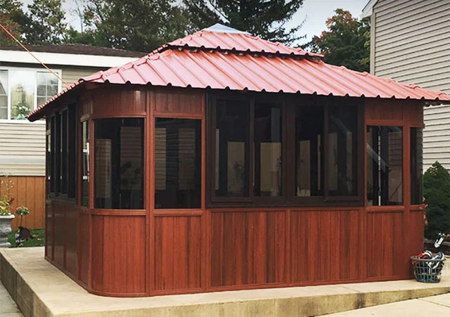 Wooden gazebo with dark windows and a red metal roof on a concrete patio.