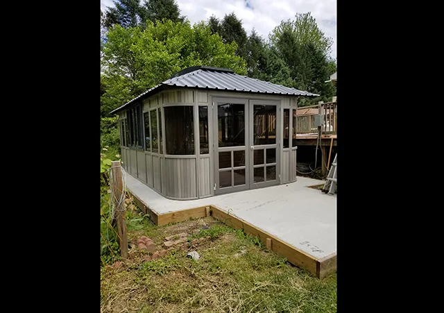 Gray metal gazebo with large windows on a concrete patio, surrounded by trees and grass.