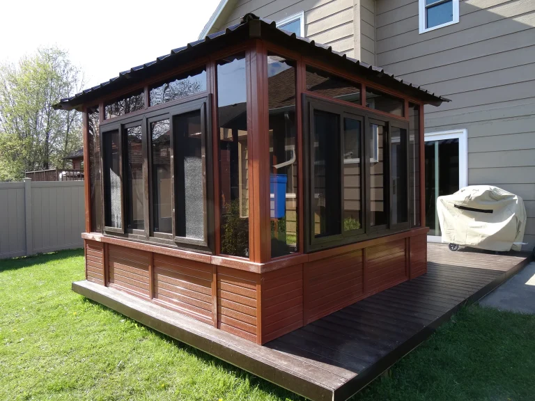Wooden sunroom with large windows and a slanted roof attached to a house, set on a deck, grass in foreground.