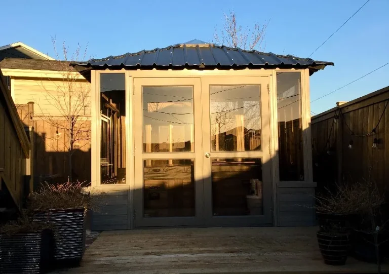 Small wooden outdoor structure with glass doors and a metal roof, surrounded by fencing and plants, in evening light.