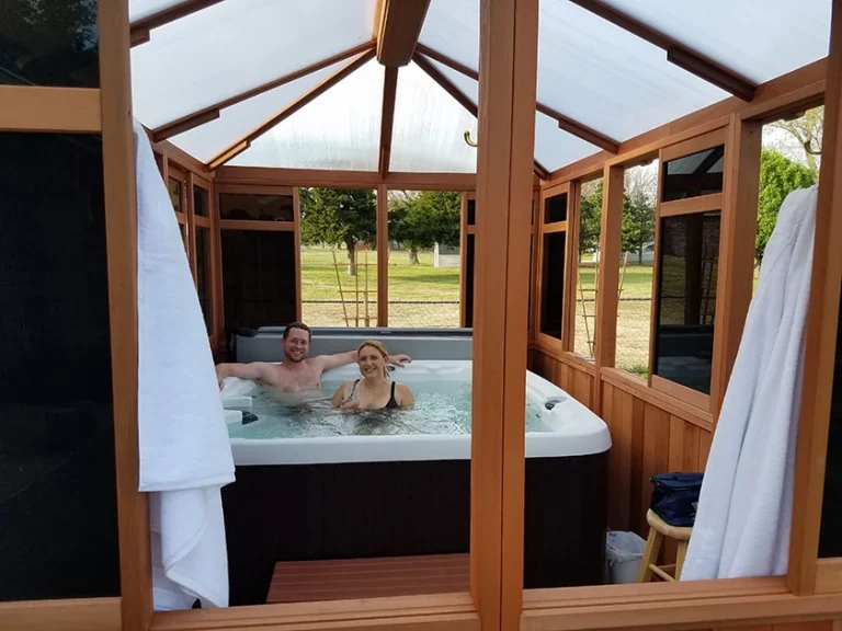 Two people relaxing in a hot tub inside a wooden enclosure with towels hanging on the sides.