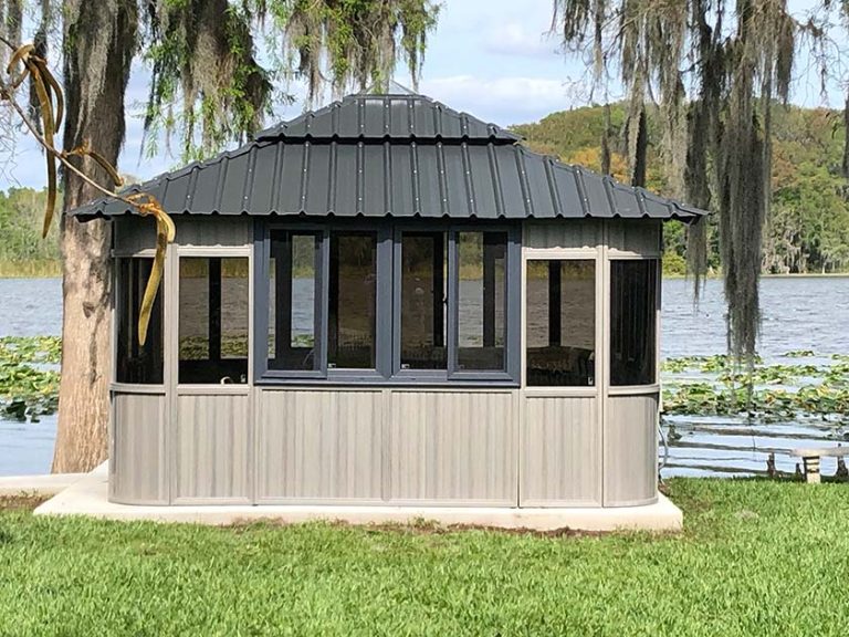 An octagonal gazebo with black roof by a lake, surrounded by trees and grass.