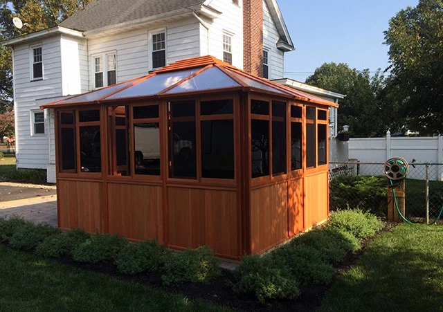 A wooden gazebo with screened windows in a backyard, near a white house with a chimney and greenery around.