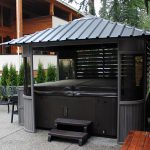Covered hot tub with steps, surrounded by greenery and patio furniture, next to a wood-paneled house.
