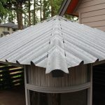 A metal roof with raindrops, atop a gazebo-like structure surrounded by trees and nearby houses.