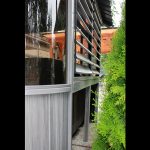Close-up of a modern building corner with slatted windows, adjacent to green foliage on a gravel surface.